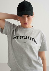 Šedé bavlněné tričko s výrazným černým nápisem "M'N' SPORTSWEAR". V kombinaci s černou kšiltovkou, která vytváří ležérní vzhled.