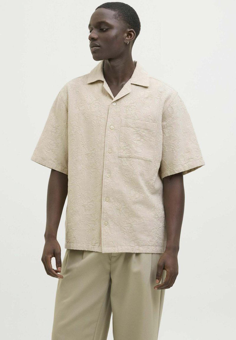 Camicia a maniche corte in tessuto beige chiaro con una sottile fantasia floreale, dotata di colletto e taschino sul petto. Abbinata a pantaloni beige.