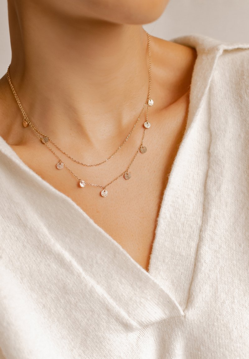 Golden layered necklace featuring small circular pendants with a textured surface, resting above a white sweater with a V-neck design.