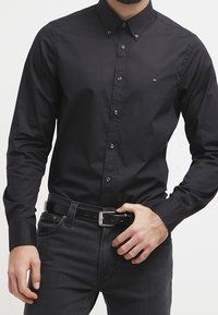 Man in een fitted zwart overhemd met een klein logo op de borst, een zwarte riem en donkere jeans die zijn riemgesp aanpast.