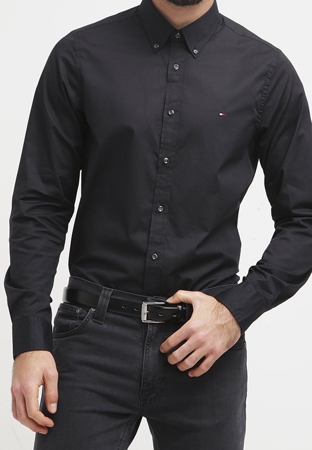 Man in een fitted zwart overhemd met een klein logo op de borst, een zwarte riem en donkere jeans die zijn riemgesp aanpast.