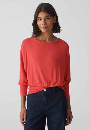 Koraalkleurige blouse met lange mouwen, ontspannen pasvorm, geplooid tailleband, ronde halslijn en een zachte brei-textuur, gecombineerd met donkere jeans.