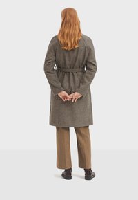 Abrigo largo a cuadros con cintura ceñida, hecho de una tela texturizada. Lleva puesto sobre pantalones marrones de pierna ancha y zapatos negros.