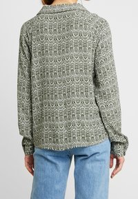 Grönmönstrad skjorta med långa ärmar, krage och ett texturerat tyg med vita blommönster. Matchad med ljusblå jeans.