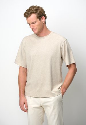 Camisa de algodón beige de manga corta, con escote redondo y un corte relajado. Se lleva con pantalones de color claro, con textura mínima.