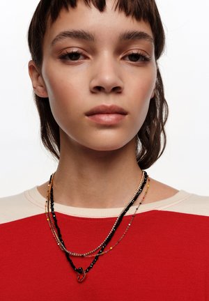 Persona joven con cabello corto y castaño que lleva collares de cuentas y cadenas superpuestos, y una camiseta roja y beige sobre un fondo blanco.