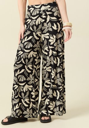 Luźne czarne spodnie z dużymi kremowymi wzorami kwiatów i liści, noszone z czarnymi sandałami na otwarte palce oraz na jednej ręce z grubymi, nałożonymi bransoletkami.