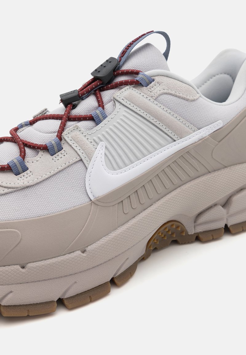 Sneaker grigio chiaro con tomaia testurizzata, lacci elastici rossi e blu, e logo Nike bianco. Presenta una suola spessa con battistrada in gomma color beige.