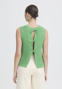 Femme aux cheveux noirs attachés en chignon, portant un haut vert en tricot sans manches noué par trois nœuds dans le dos, associé à un pantalon crème taille haute.