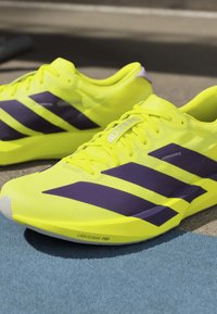 Φωτεινά κίτρινα αθλητικά παπούτσια με μαύρες ρίγες και σήμανση "ADIZERO", ακουμπούν σε μπλε χαλί και γκρι επιφάνεια.