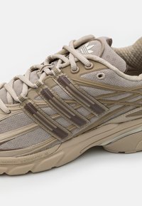 Beige idrottssko med mesh-överdel, med tre bruna plastremsor och en strukturerad gummisula. Snören är beige och med en vadderad krage.