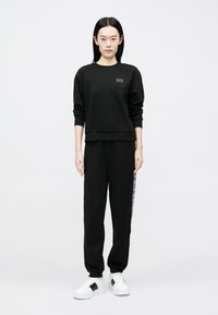 Mladá žena oblečená v čiernej mikine EA7 a zodpovedajúcich čiernych teplákoch s logom na boku, stojaca pred jednoliatym bielym pozadím.