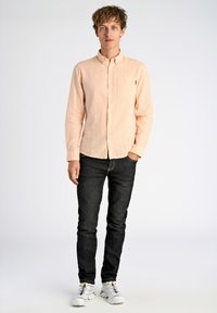 Helles Pfirsichfarbenes Hemd mit langen Ärmeln und einer Brusttasche, kombiniert mit schwarzen Jeans und weißen Turnschuhen.