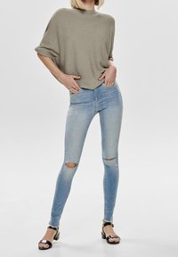 Femme portant un haut ample vert olive, un jean skinny bleu clair déchiré et des sandales noires à talons carrés et ouverts, debout les mains dans les poches.