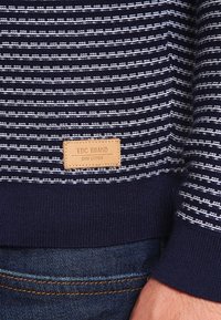 Laivastonsininen ja valkoinen raidallinen neule, jossa on resorireuna ja edessä alaosassa nahkainen merkki, jossa lukee "EDC BRAND DNM GOODS".