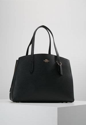 Zwarte leren Coach-tas met dubbele handvatten, gouden logo en een klein leren hangertje, geplaatst op een wit voetstuk.