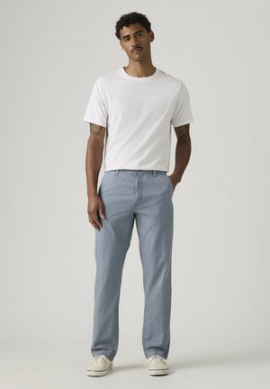 Man with curly hair wearing a white short-sleeve t-shirt, light blue pants, and white sneakers, standing with one hand in pocket.