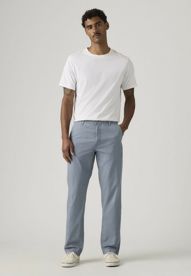 Mand med krøllet hår iført en hvid t-shirt med korte ærmer, lyseblå bukser og hvide sneakers, stående med den ene hånd i lommen.