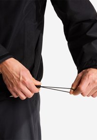 Des mains ajustant le cordon de serrage sur l'ourlet d'une veste noire sur un fond blanc uni.
