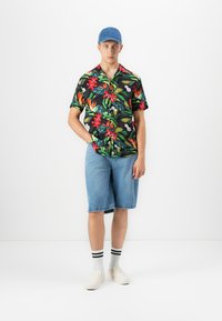 Jonge man met een blauwe pet, een zwart bloemenoverhemden met korte mouwen, denim shorts tot net boven de knie, witte sokken met zwarte strepen en witte instapschoenen.