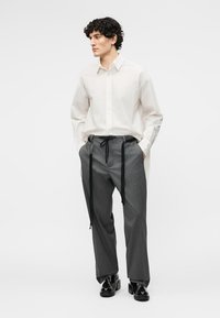 Mann trägt ein weißes, langärmliges Hemd mit Knöpfen, graue Nadelstreifenhose mit langen schwarzen Kordeln und schwarze glänzende Anzugschuhe.