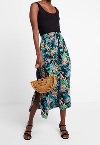 Gonna maxi floreale con motivi verdi, rosa e blu, caratterizzata da uno spacco laterale. Abbinata a un top nero e a una borsa in bambù.
