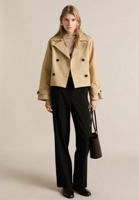 Beige cropped double-breasted jacket with black buttons, paired with black wide-leg pants and a tan knit top, holding a black handbag.