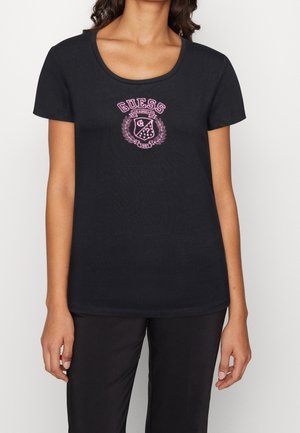 Woman wearing a black short-sleeve t-shirt with a pink Guess logo and crest in the center front, paired with black pants.