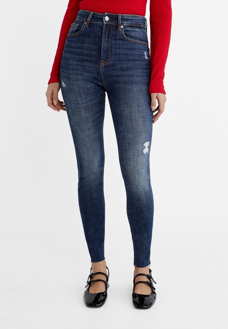 Smala blå jeans med hög midja, med ett bleknat märke och subtila nötningsmärken på högra låret. Kompletterat med svarta lackskor.