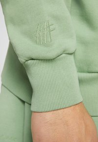 Gros plan sur un poignet de sweat-shirt côtelé vert clair avec un logo brodé discret sur la manche au-dessus du poignet d'une personne.