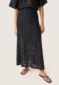 Jupe maxi en dentelle noire avec un motif floral, dotée d'une taille cintrée et d'un ourlet qui révèle un peu de peau en dessous. Associée à des tongs noires.