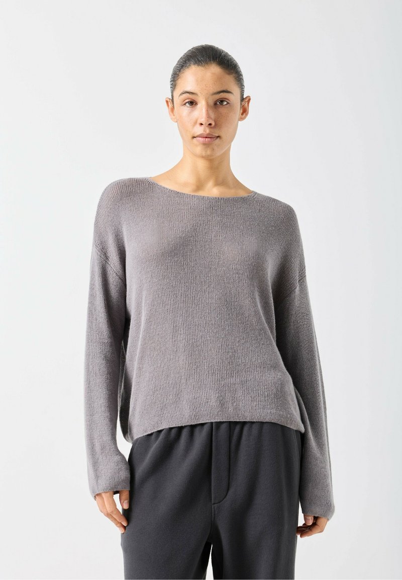 Gaiši pelēks adīts džemperis ar plašu kakla izgriezumu un garām piedurknēm, ar brīvu piegriezumu un fakturētu virsmu, valkāts kopā ar tumšām biksēm.