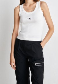 Vit ribbad linne med rund halsringning som har en liten svart CK-logotyp på bröstet; bärs med svarta byxor med en dragkedjeficka.