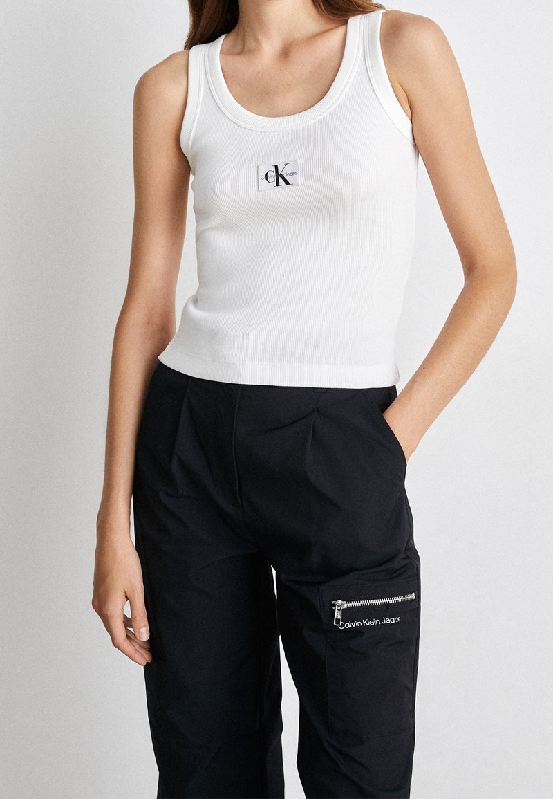 Vit ribbad linne med rund halsringning som har en liten svart CK-logotyp på bröstet; bärs med svarta byxor med en dragkedjeficka.