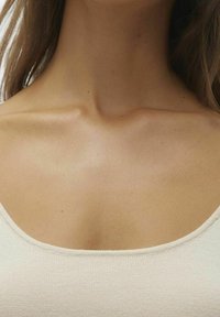 Beige stickad tyg topp med rundad halsringning, med en mjuk textur och subtila detaljer längs kanterna, som framhäver nyckelbenet.