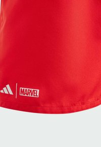 Pantalones cortos deportivos rojos hechos de un tejido suave, con los logotipos blancos de Adidas y Marvel cerca del dobladillo. El diseño es minimalista, sin patrones.