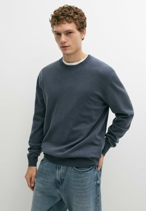 Jeune homme aux cheveux bouclés portant un pull bleu foncé à col rond par-dessus une chemise blanche et un jean bleu clair, debout devant un fond blanc uni.