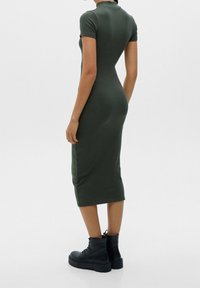 Oliivinvihreä vartalonmyötäinen mekko, jossa on lyhyet hihat, korkea kaulus ja midi-pituus. Sileä, joustava kangas, jossa sivuhalkiot. Yhdistetty mustiin korokepohjasaappaisiin.