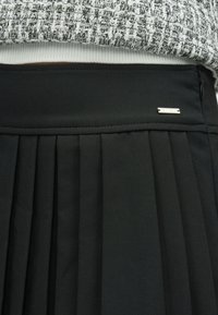 Zwarte geplooide rok met een gladde tailleband en een kleine zilveren accenten. De stof heeft een zachte textuur en een minimalistisch ontwerp.