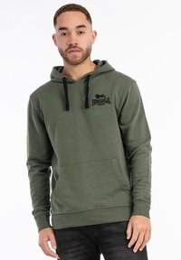 Sudadera de color verde oliva hecha de una tela suave, con cordones negros, un bolsillo tipo canguro y un logo en el lado izquierdo del pecho.