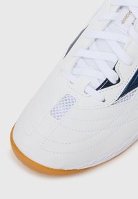 Sapatilha atlética branca com um cabedal sintético suave, apresentando detalhes em azul, malha e uma sola de borracha texturizada de cor caramelo.