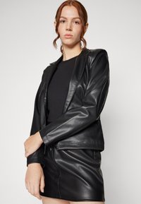 Veste en cuir noir au design ajusté, avec un col cranté et des manches longues. Associée à une mini-jupe en cuir noir assortie. Texture lisse.