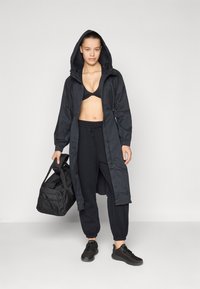 Black longline rain jacket with hood, worn over a black sports bra. Paired with black joggers and black sneakers, holding a black duffel bag.