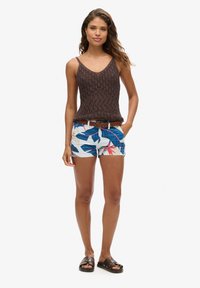 Top de punto marrón, shorts blancos con un patrón de hojas azules y acentos rojos, cinturón trenzado marrón y sandalias marrones oscuras.