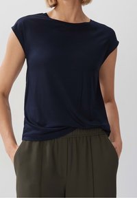 Námořnická modrá halenka s krátkým rukávem vyrobená z hladké látky s volným střihem. Má kulatý výstřih a jemně se skládá přes olivově zelené kalhoty.