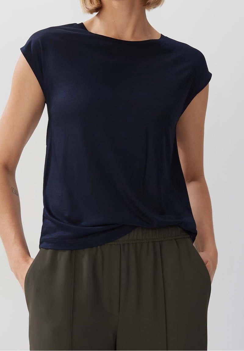 Námořnická modrá halenka s krátkým rukávem vyrobená z hladké látky s volným střihem. Má kulatý výstřih a jemně se skládá přes olivově zelené kalhoty.