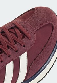 Gros plan d'une basket bordeaux avec une tige en suède et tissu, des bandes blanches sur le côté, des lacets bordeaux et une semelle blanche avec un liseré bleu foncé.