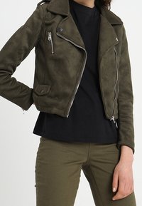 Donna che indossa una giacca biker in suede verde oliva sopra una maglietta nera e pantaloni verde oliva, in posa con una mano sui fianchi.