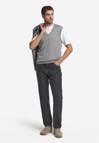 Grå ullväst över en vit T-shirt, svarta jeansbyxor och beiga sneakers. Personen håller en grå vadderad jacka. Neutralt bakgrund.