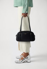 Bolso negro con dobles asas, logo dorado, diseño elegante, textura suave, combinado con pantalones crema y tacones plateados metálicos.